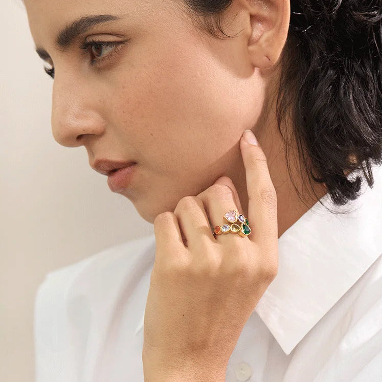 A gold-tone wrap ring featuring a bypass design set with multi-coloured heart-shaped crystals, including pink, purple, emerald green, and amber tones.